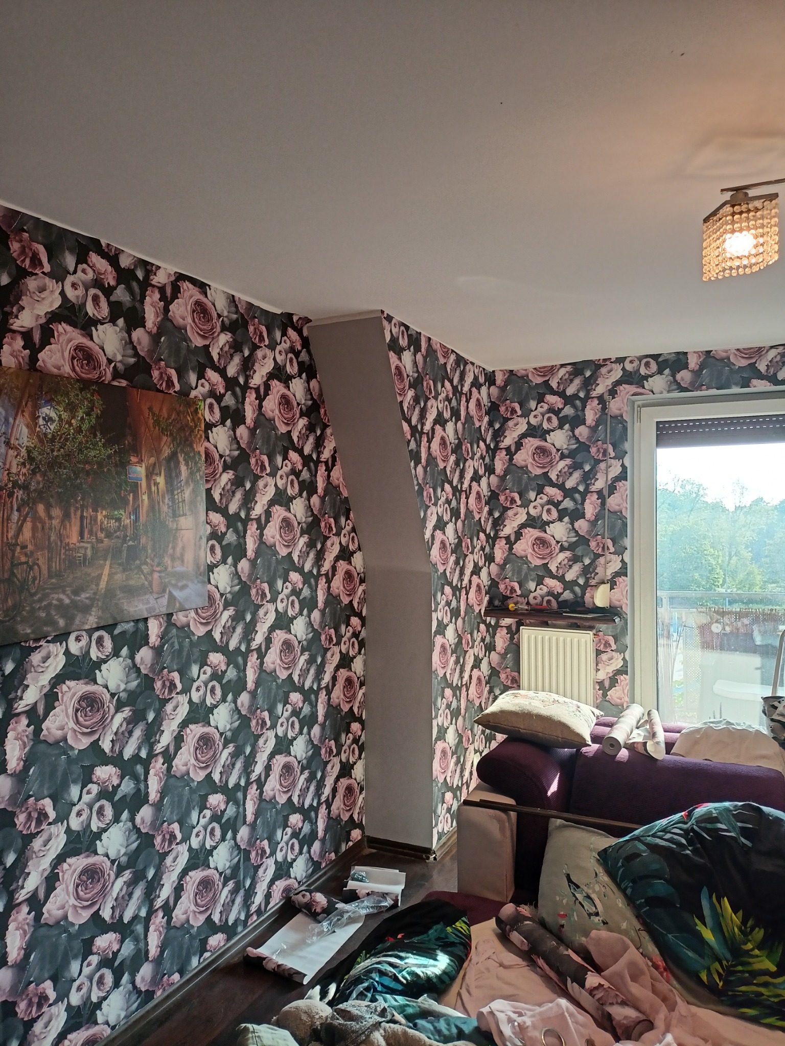 Pokój z tapetą w różowe róże, widoczny fragment sofy, okno i obraz na ścianie.