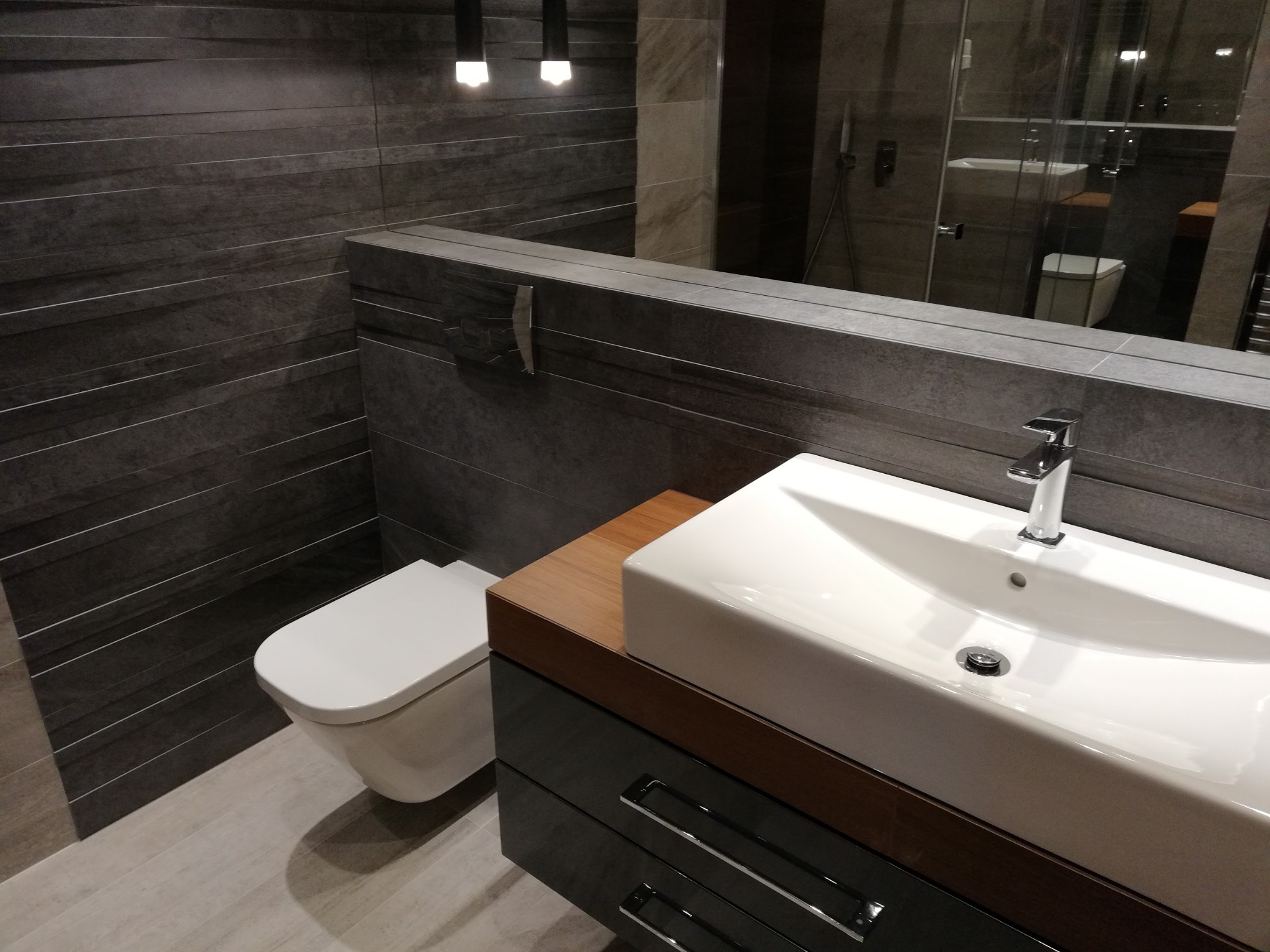 Nowoczesna łazienka z umywalką nablatową, podwieszaną toaletą i szarą, poziomą okładziną ścienną, odbicie kabiny prysznicowej w lustrze.