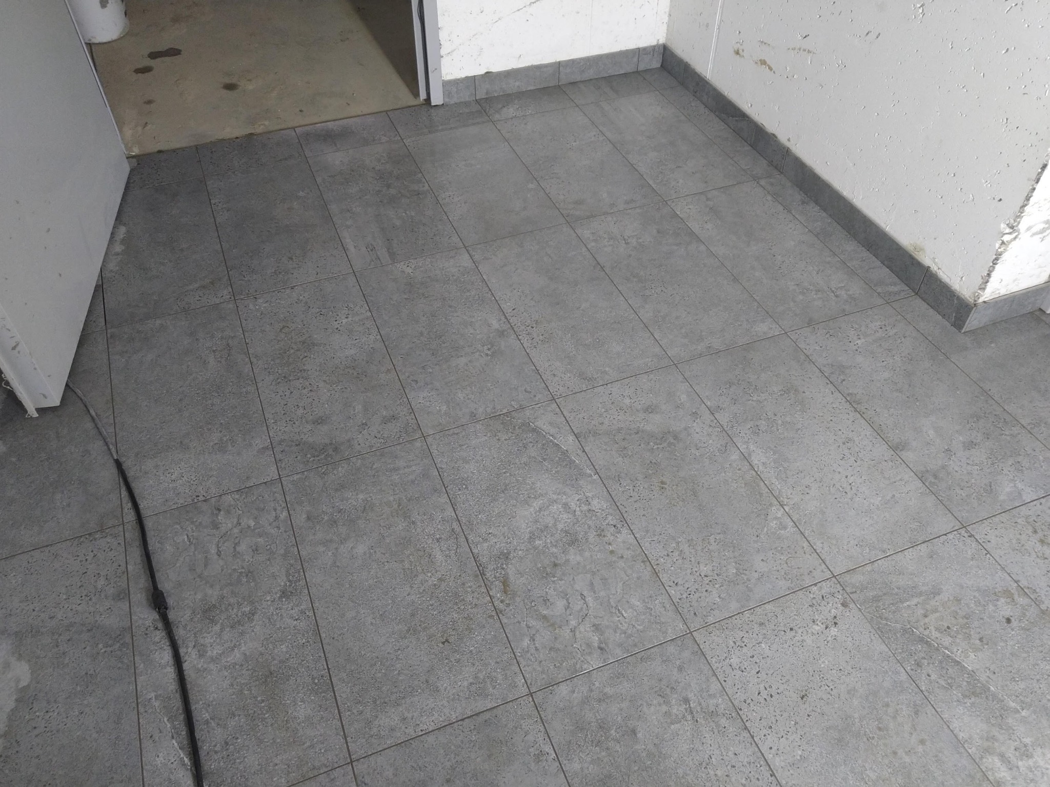 Ułożona szara podłoga z płytek ceramicznych w pomieszczeniu, widoczne płytki przypodłogowe i fragment ściany z odpadającym tynkiem.