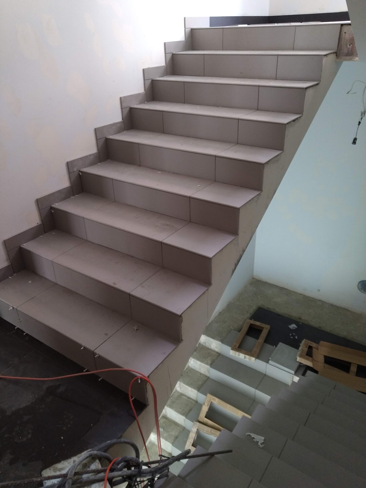 Schody wyłożone jasnymi płytkami ceramicznymi w trakcie montażu; widoczne krzyżyki dystansowe, przewód elektryczny i narzędzia na dole kadru.