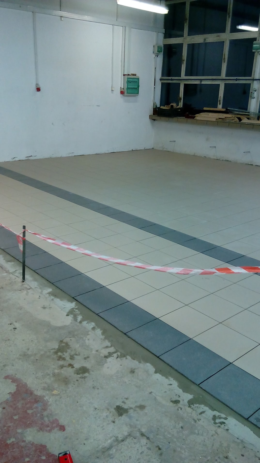 Wnętrze pomieszczenia z nową, jasną podłogą wyłożoną płytkami w szachownicę z ciemnoszarymi pasami, zabezpieczone taśmą ostrzegawczą na metalowym słupku, widoczne ślady wilgoci i zabrudzeń...