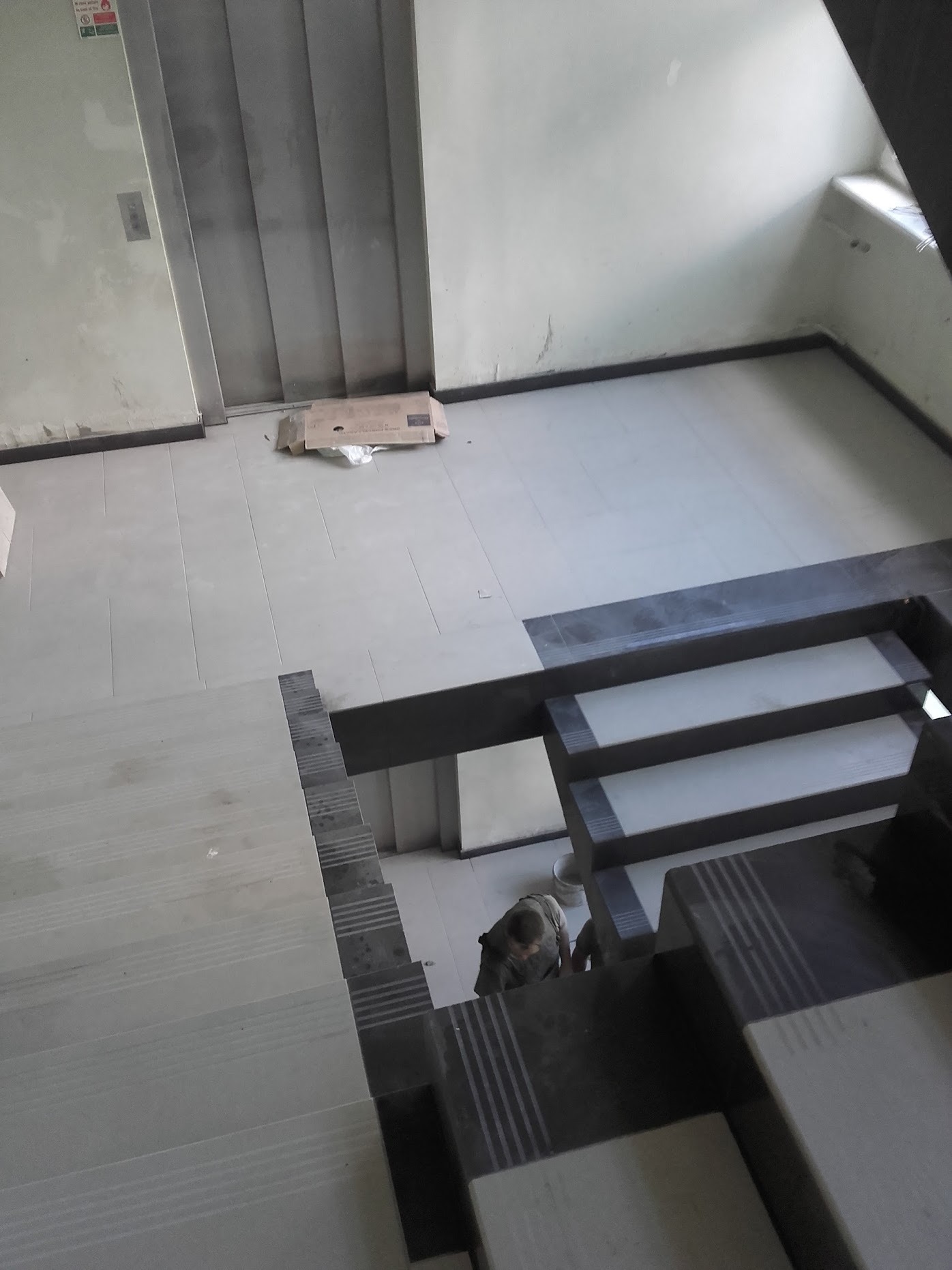 Widok z góry na klatkę schodową z wyłożonymi płytkami stopnicami w odcieniach szarości, w tle jasna podłoga i ściany, przy windzie leży karton, osoba w trakcie pracy na schodach.