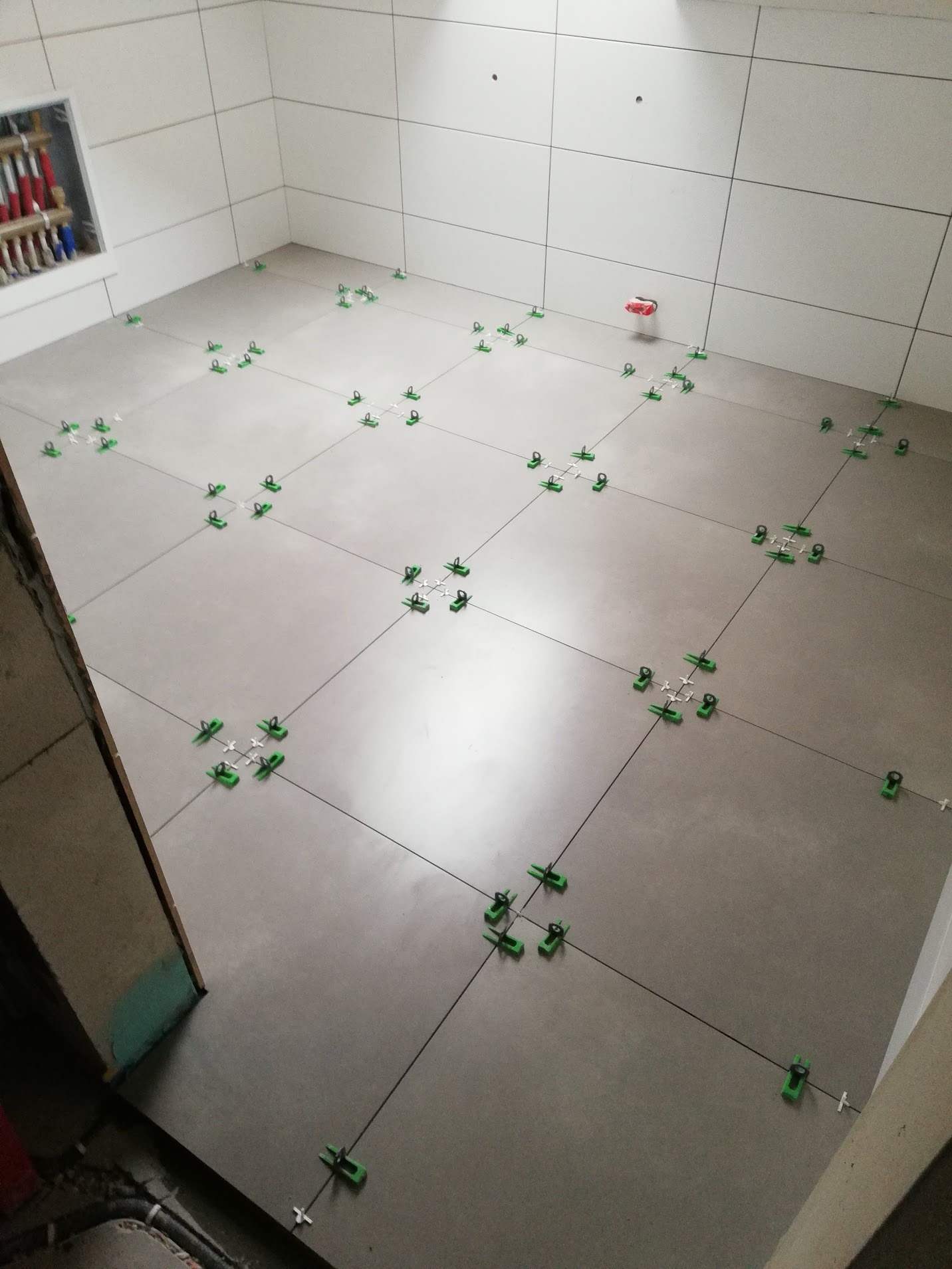 Szare płytki podłogowe w trakcie układania, z systemem poziomowania płytek (klipsy i kliny) w kolorze zielonym, widoczne także białe płytki ścienne i fragment instalacji wodnej w rogu pomieszczenia.