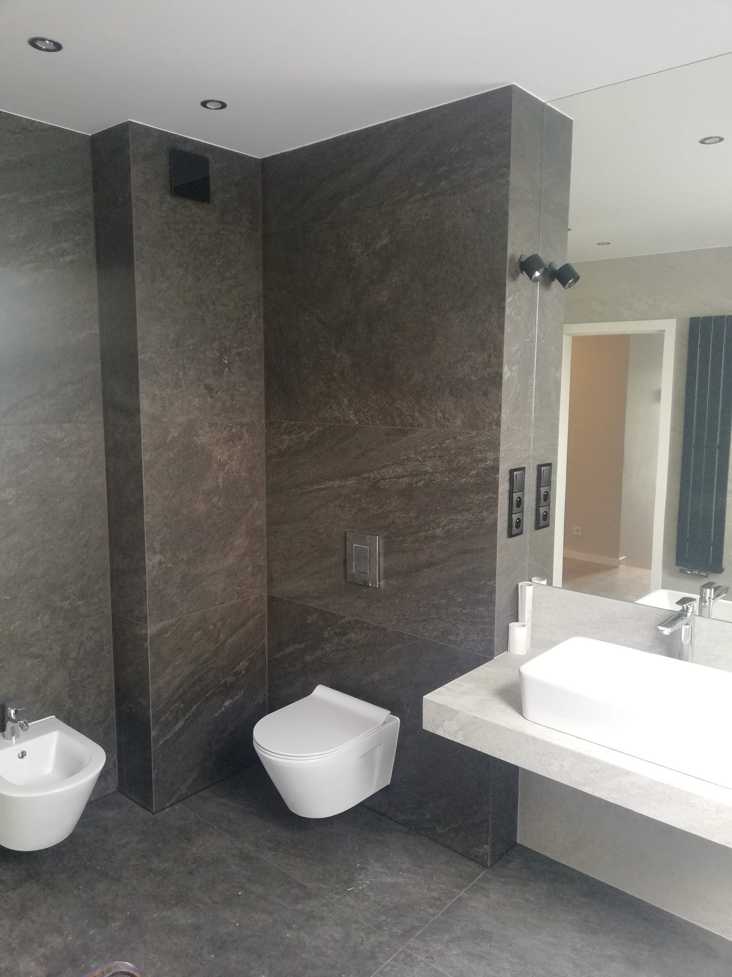 Nowoczesna łazienka z szarymi płytkami imitującymi kamień, wiszącą toaletą i bidetem, minimalistyczną umywalką nablatową oraz dużym lustrem z oświetleniem punktowym.