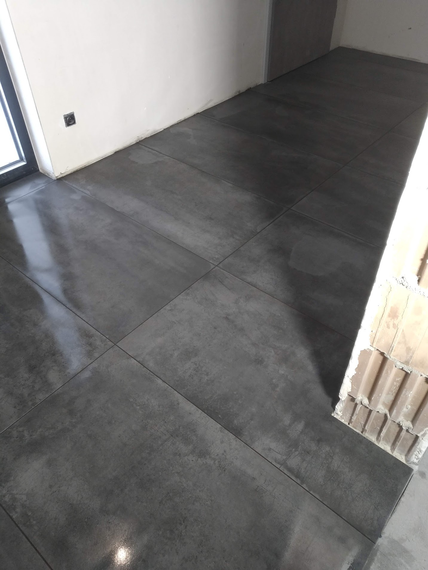 Ułożone, ciemnoszare płytki podłogowe w minimalistycznym wnętrzu, z widoczną instalacją elektryczną w ścianie i fragmentem ściany z cegieł.