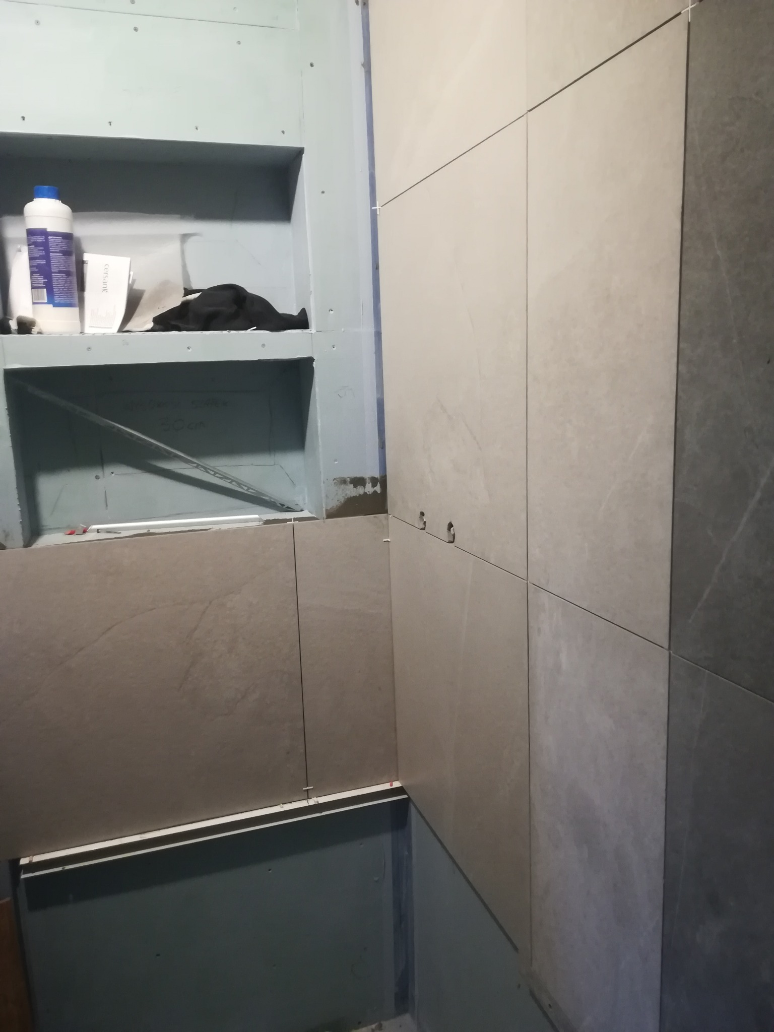 Układanie płytek w łazience, widoczna zabudowa z półkami z płyty gipsowej, częściowo wykończona ścianka z jasnoszarych płytek z krzyżykami dystansowymi, przygotowane otwory na instalację.