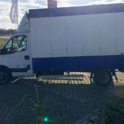 Biały samochód dostawczy z plandeką, zaparkowany na ażurowym parkingu, widok z boku, z widocznym logo firmy transportowej na drzwiach kabiny.