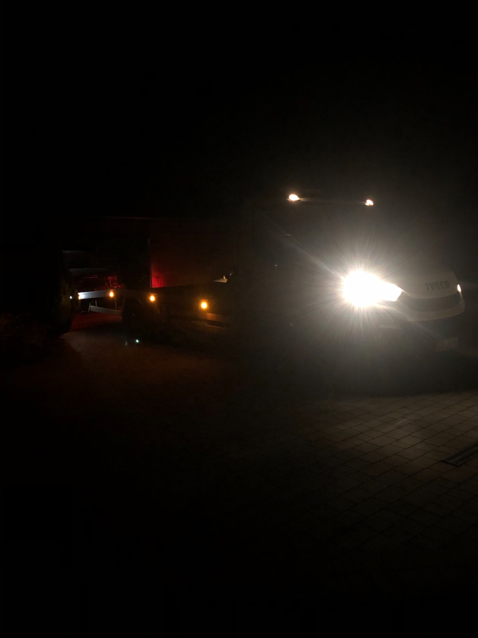 Ciężarówka marki Iveco z lawetą w ciemności, oświetlona reflektorami, widoczne światła pozycyjne lawety.
