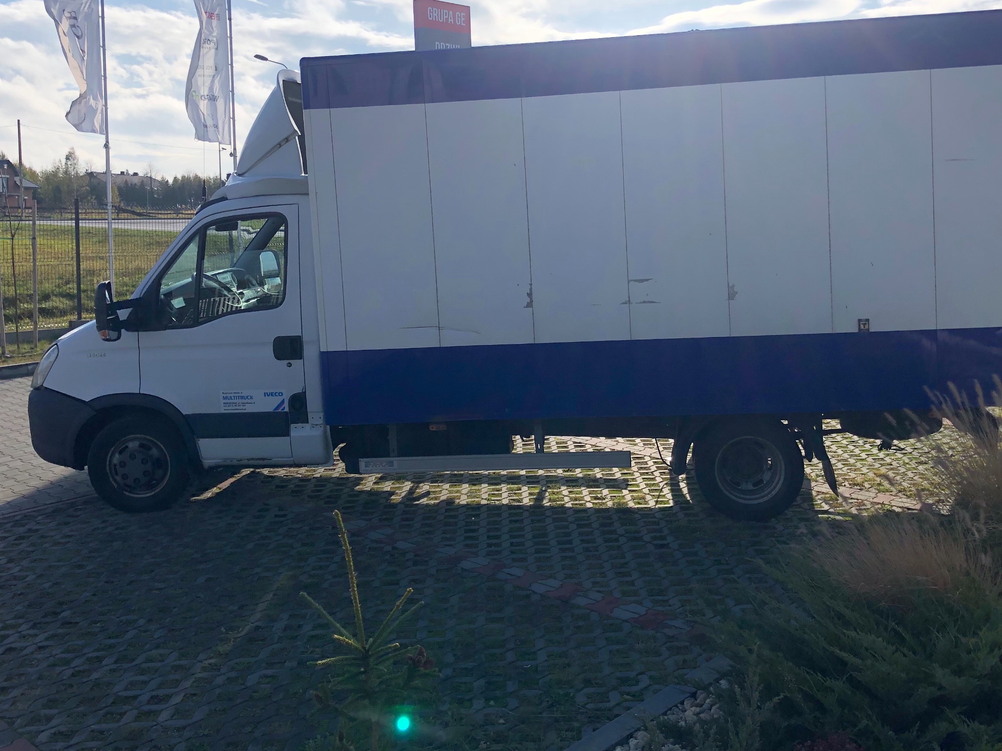 Biały samochód dostawczy z plandeką, zaparkowany na ażurowym parkingu, widok z boku, z widocznym logo firmy transportowej na drzwiach kabiny.