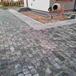 Ułożona kostka brukowa w odcieniach szarości prowadząca do domu z widoczną betoniarką i paletą z kostką obok, w tle trawnik i drzewa.
