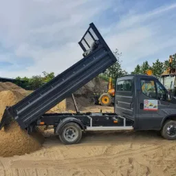 Wywrotka z podniesioną skrzynią kipruje piasek na ziemię. W tle koparki i hałda piasku. Szare auto z logo firmy. Błękitne niebo z chmurami.