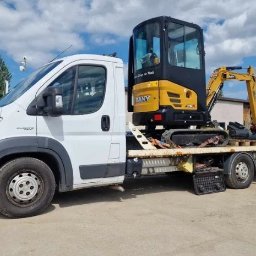 Mini Budowlanka - Żółta minikoparka SANY na lawecie białego samochodu dostawczego, gotowa do transportu. Na drugim planie widoczny teren budowy z koparką.