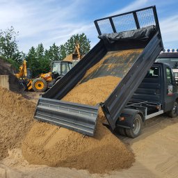 Mini Budowlanka - Wywrotka w trakcie wysypywania piasku na placu budowy; w tle koparka JCB i ciężarówka, błękitne niebo.