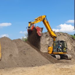 Mini Budowlanka - Żółta koparka z przesiewaczem nabiera czarnoziem na pryzmę. Błękitne niebo z chmurami w tle. Widoczny operator w kabinie maszyny. Przesiewanie ziemi.