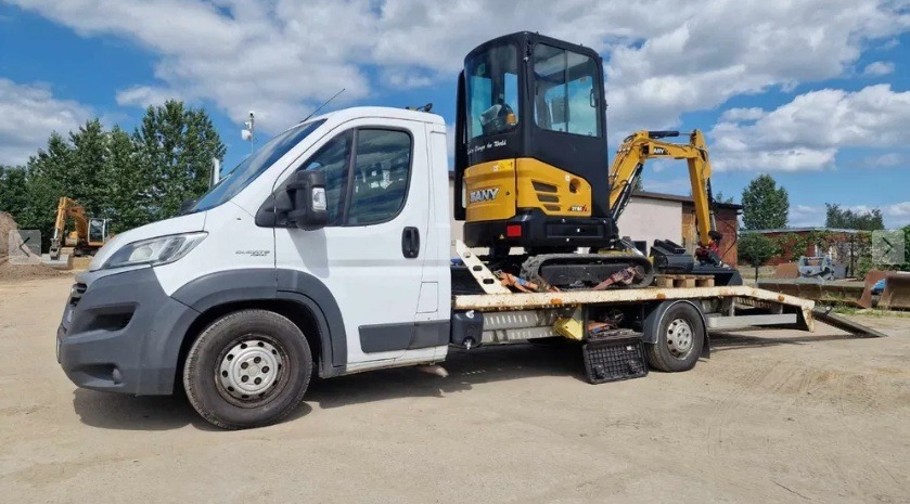 Żółta minikoparka SANY na lawecie białego samochodu dostawczego, gotowa do transportu. Na drugim planie widoczny teren budowy z koparką.