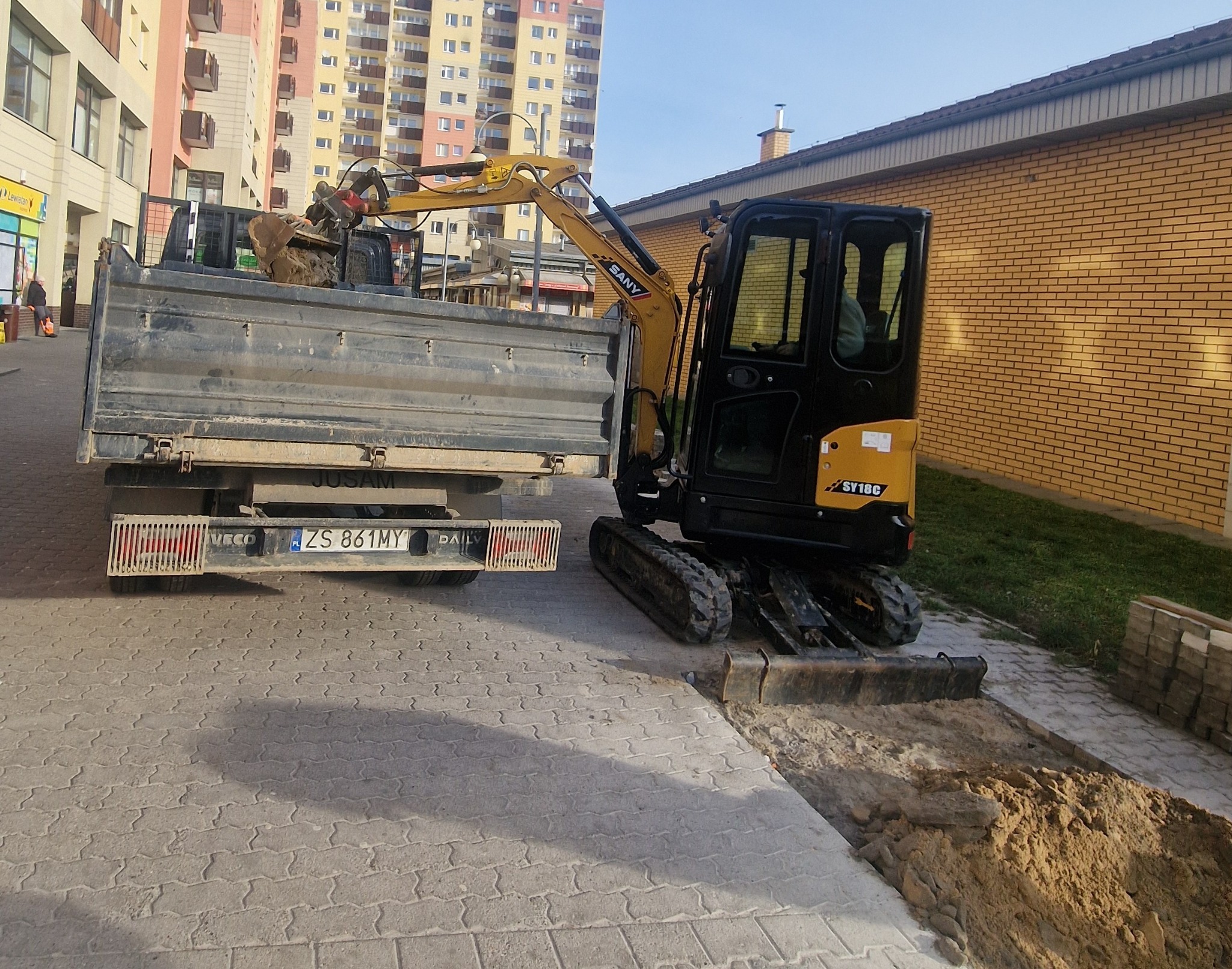 Żółta minikoparka SANY ładuje ziemię do skrzyni ciężarówki na tle miejskiej zabudowy. Widoczny wykop w kostce brukowej i sterta ziemi w skrzyni ładunkowej.