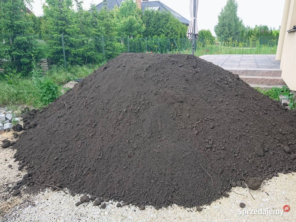 Kupa ciemnej ziemi na jasnym podłożu, w tle zielona roślinność i fragment domu z tarasem. Ziemia usypana w stożek, widoczne grudki i nierówności.