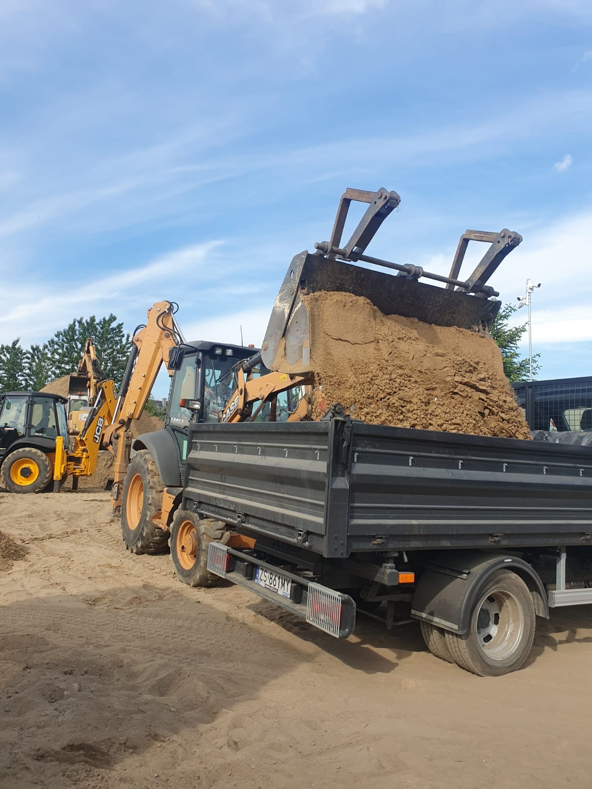 Koparko-ładowarka CASE ładuje piasek do wywrotki na placu budowy; w tle druga maszyna, błękitne niebo.