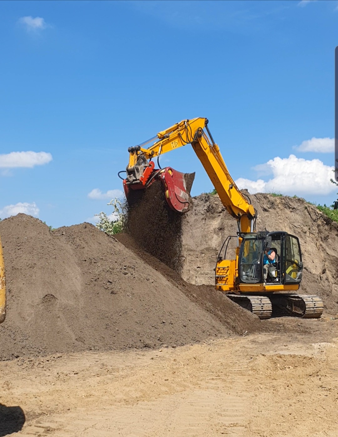 Żółta koparka z przesiewaczem nabiera czarnoziem na pryzmę. Błękitne niebo z chmurami w tle. Widoczny operator w kabinie maszyny. Przesiewanie ziemi.