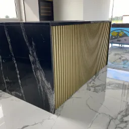 Czarny blat z marmurowym wzorem połączony z pionowymi, złotymi panelami dekoracyjnymi, całość na białej podłodze z szarymi żyłami, widoczne odbicie w polerowanej powierzchni.