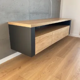 Wisząca szafka RTV z dwiema szufladami o froncie w kolorze jasnego drewna i czarnych bokach, zamontowana na ścianie z szarych płyt betonowych, oświetlona lampą wiszącą.