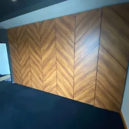 Drewniana ściana z paneli ułożonych w jodełkę, o ciepłym odcieniu brązu, na tle ciemnej podłogi i sufitu, oświetlona punktowo.