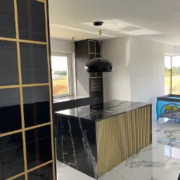 Nowoczesna kuchnia z wyspą o czarnym marmurowym blacie i złotymi pionowymi listwami, czarne szafki z złotymi ramkami, podłoga z białego marmuru, czarna lampa wisząca, widok z okna na pole.