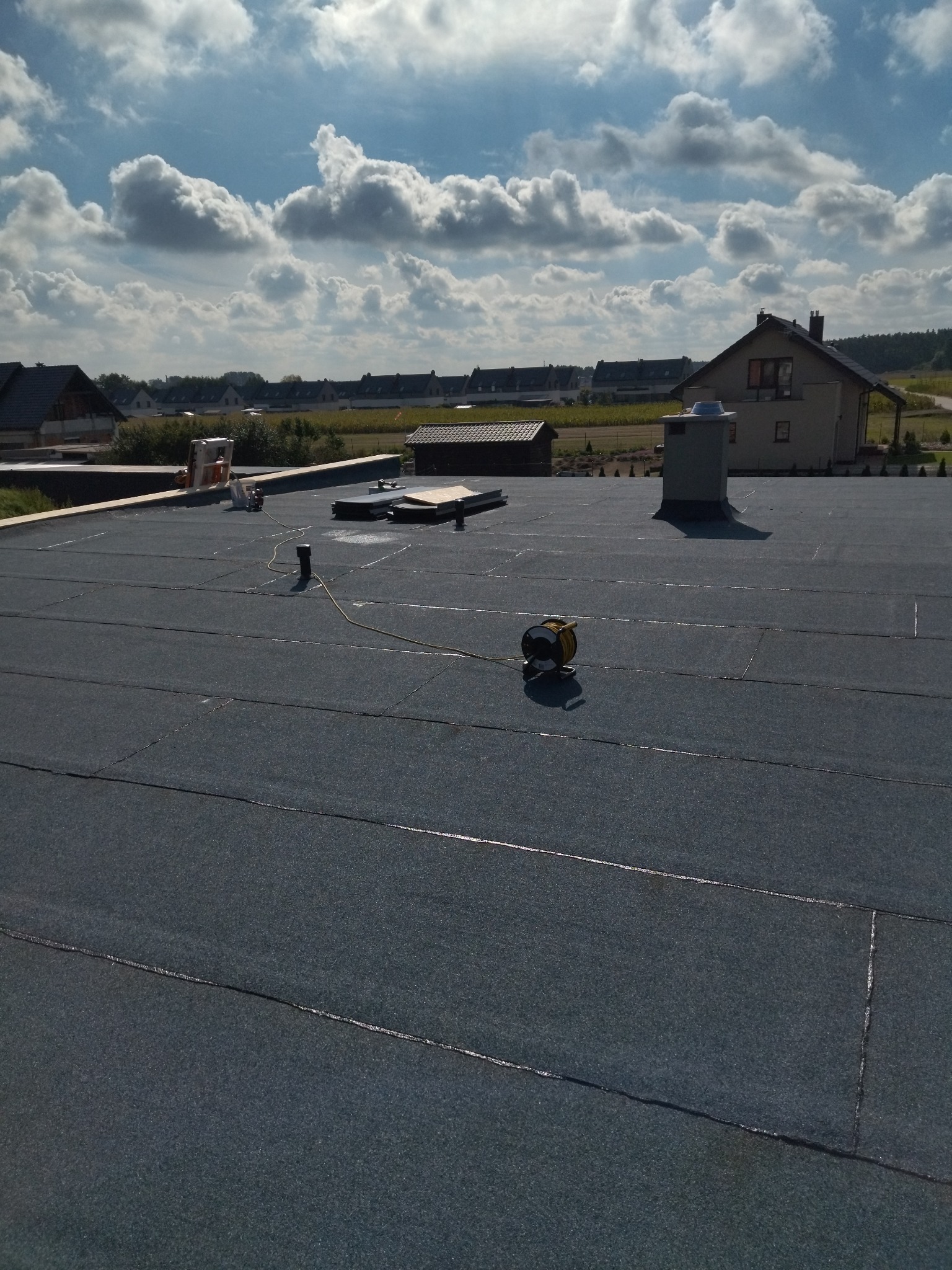 Płaski dach pokryty papą termozgrzewalną z widocznym kominem, zwiniętym przedłużaczem i elementami wentylacji, na tle zabudowy jednorodzinnej i błękitnego nieba z chmurami.