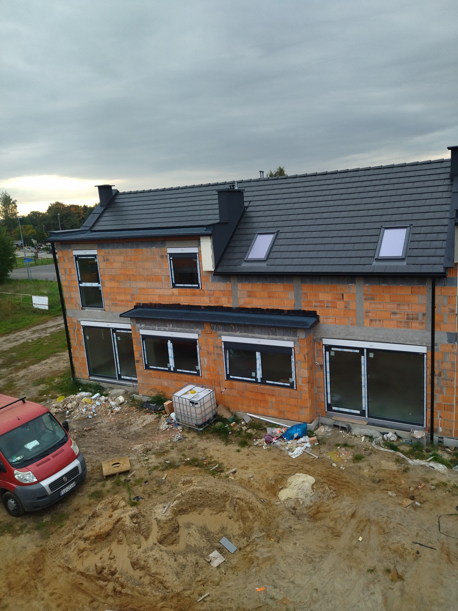 Wysoki kąt na dwupiętrowy dom w trakcie budowy z ciemnoszarym dachem, widoczne okna dachowe, czerwona cegła, częściowo zamontowane okna i czerwony samochód dostawczy w pobliżu.