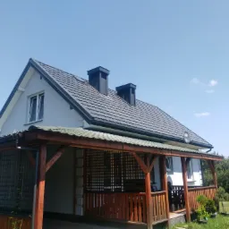 Dom jednorodzinny z szarą dachówką, drewnianym gankiem i dwoma czarnymi kominami na tle błękitnego nieba.