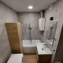 Ultimum Wykończenia - Wykończona łazienka z szarymi płytkami, drewnianą szafką pod umywalką i wysoką szafką obok wanny, widoczny bojler i pralka, oświetlenie sufitowe.