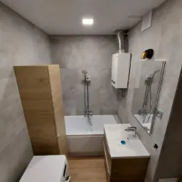 Wykończona łazienka z szarymi płytkami, drewnianą szafką pod umywalką i wysoką szafką obok wanny, widoczny bojler i pralka, oświetlenie sufitowe.