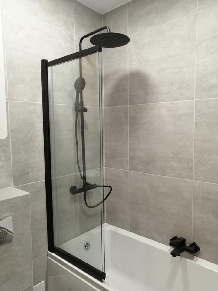 Nowoczesna kabina prysznicowa z czarnymi elementami w łazience wyłożonej jasnymi, kwadratowymi płytkami. Widoczna biała wanna i bateria. Minimalistyczny design.