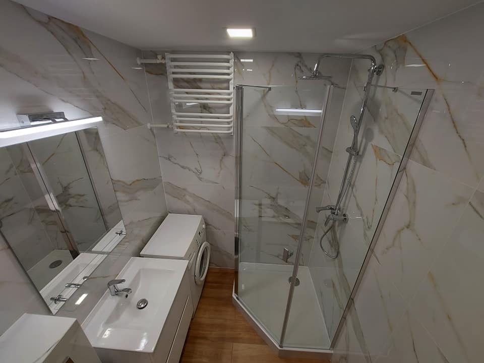 Nowoczesna łazienka z marmurowymi płytkami, kabiną prysznicową narożną, umywalką z szafką, pralką i grzejnikiem drabinkowym.