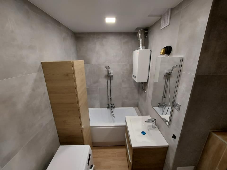 Wykończona łazienka z szarymi płytkami, drewnianą szafką pod umywalką i wysoką szafką obok wanny, widoczny bojler i pralka, oświetlenie sufitowe.