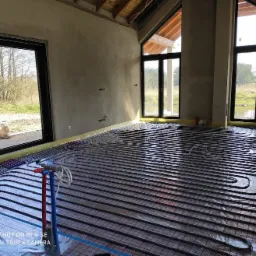 Montaż ogrzewania podłogowego w surowym wnętrzu z dużymi oknami; rury grzewcze ułożone na folii izolacyjnej, widoczne przyłącza rur czerwone i niebieskie.