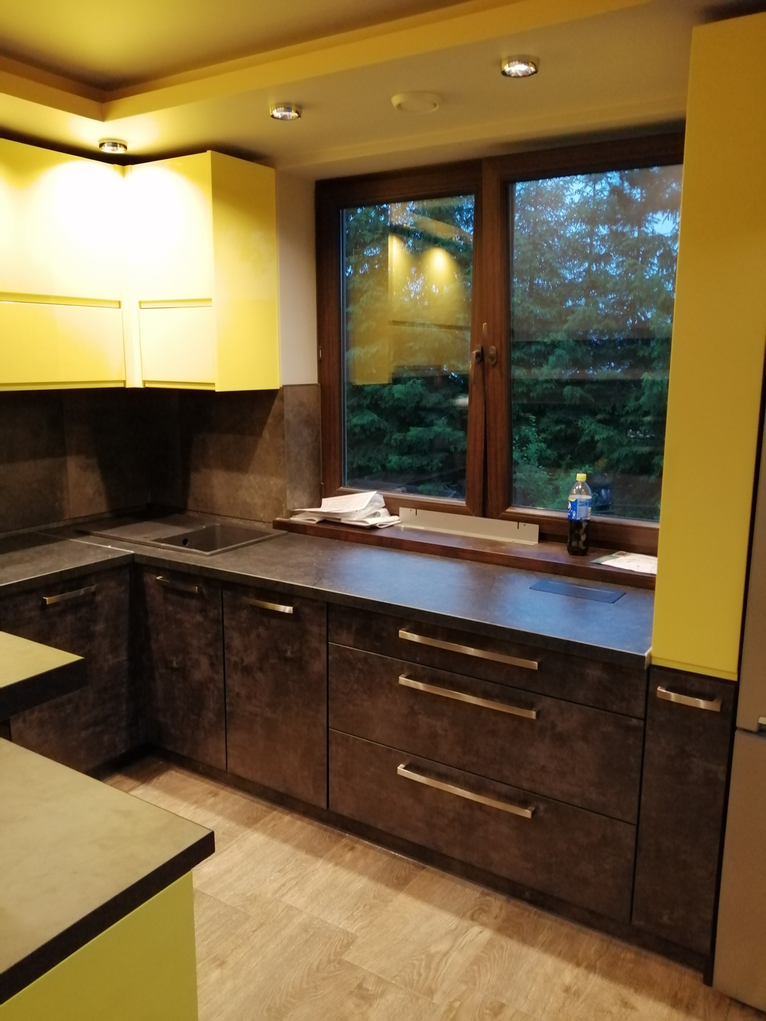 Nowoczesna kuchnia z żółtymi górnymi szafkami i ciemnobrązowymi dolnymi szafkami z poziomymi uchwytami, widok na zieleń za oknem.