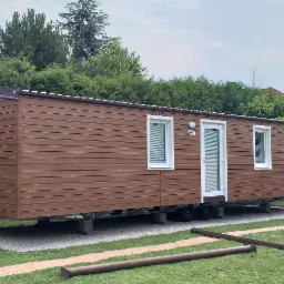 Brunatny budynek modułowy z trzema oknami i drzwiami, ustawiony na betonowych podstawach na żwirowym podłożu, z mężczyzną na kolanach przy trawniku, w tle ogrodzenie i dom.