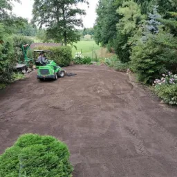Zielony traktor wyrównuje teren pod przyszły ogród, widoczne ślady opon na świeżo przekopanej ziemi, w tle drzewa i krzewy.