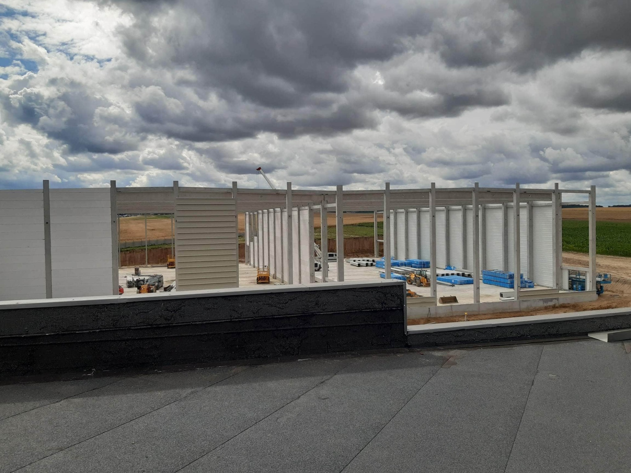 Widok z dachu płaskiego na szkielet powstającego budynku przemysłowego, z widocznymi prefabrykatami i sprzętem budowlanym na placu budowy w tle, pod pochmurnym niebem.