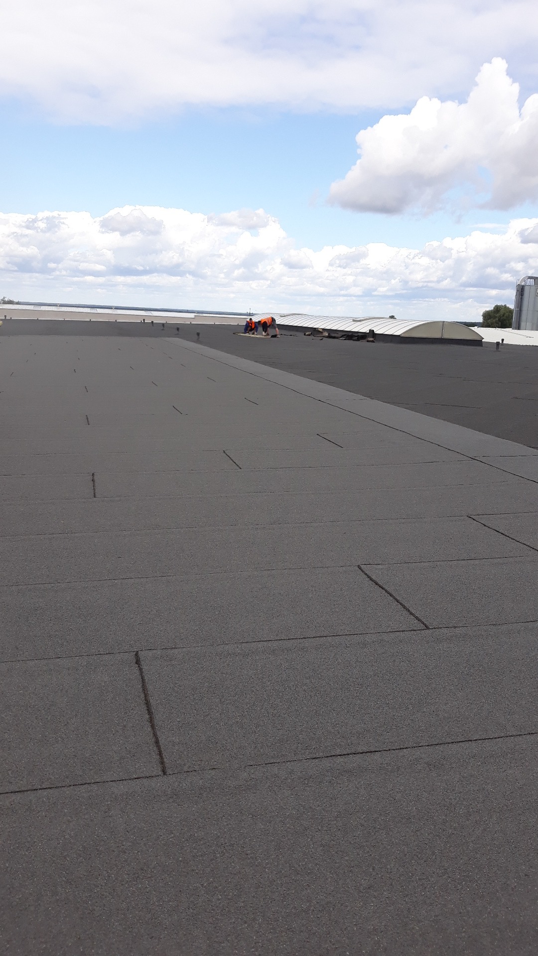 Ekipa dekarzy w pomarańczowych kamizelkach pracuje na rozległym, płaskim dachu pokrytym papą, z widocznymi łączeniami, w tle błękitne niebo z białymi chmurami i elementy konstrukcji budynku.