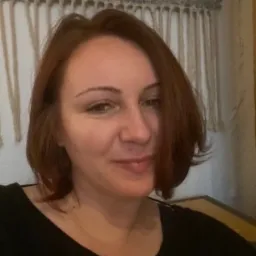 Portret kobiety z rudymi włosami, uśmiechającej się do kamery, z dekoracyjną makramą w tle.