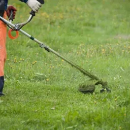 Pracownik w pomarańczowych spodniach używa podkaszarki spalinowej do trawy na łące z żółtymi kwiatami, w tle rozproszony krajobraz.
