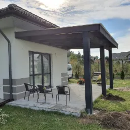 Zadaszony taras z ciemnego drewna przylegający do białego domu z szarymi akcentami, widoczne trzy czarne krzesła na jasnej kostce brukowej, trawnik z kopcami ziemi w tle.