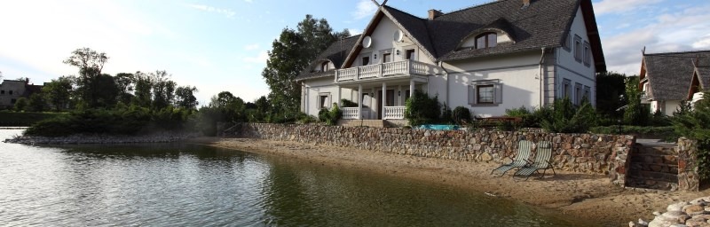 Widok na biały budynek z balkonem i dwoma krzesłami plażowymi na piaszczystym brzegu jeziora, otoczony kamiennym murem i zielenią.