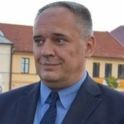 Portret mężczyzny w ciemnym garniturze i niebieskiej koszuli z krawatem na tle budynku z czerwoną dachówką.