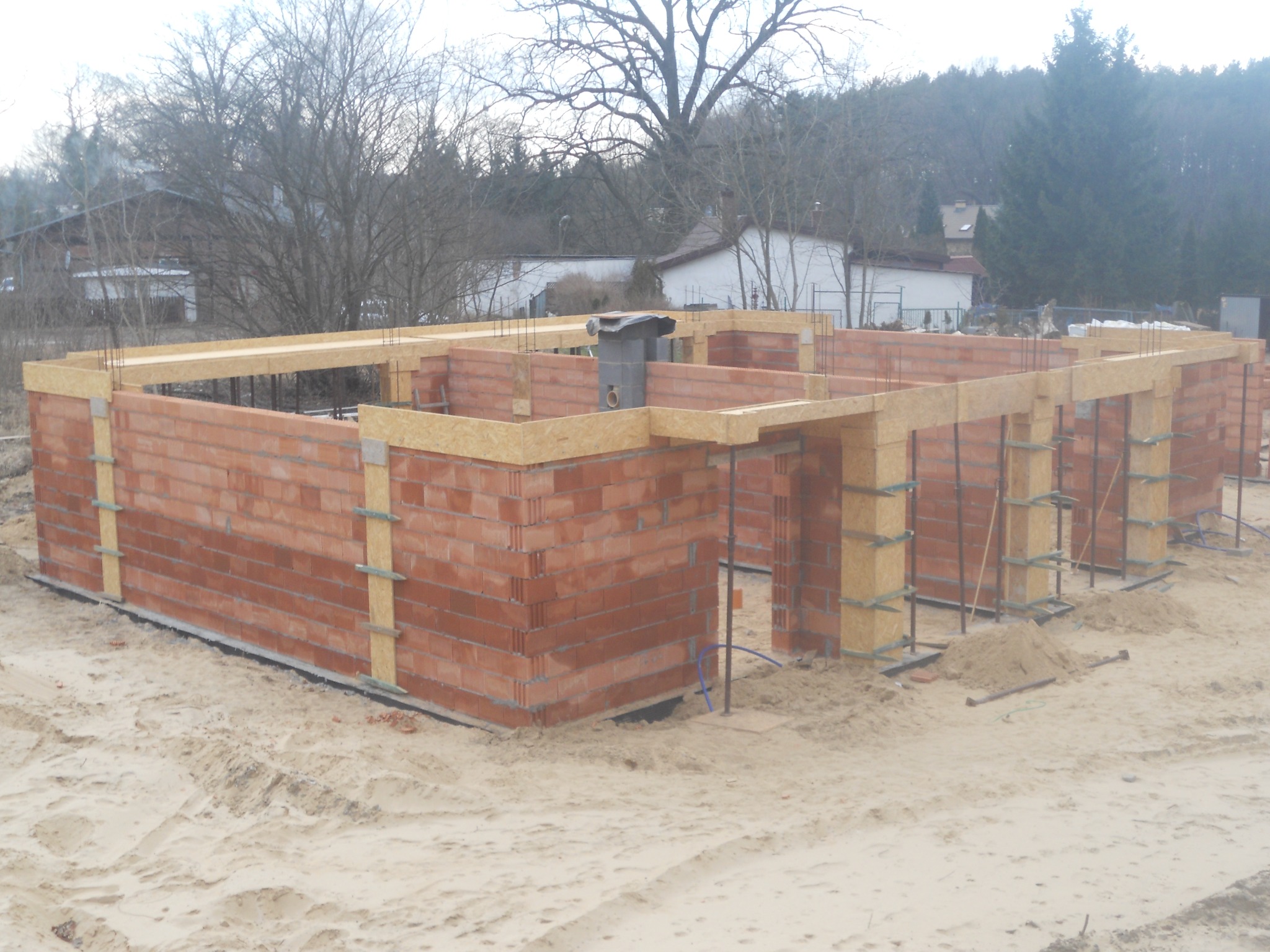 Wznoszone mury parteru domu z czerwonej cegły, z widocznymi elementami szalunku i zbrojenia, na piaszczystym podłożu w otoczeniu drzew i budynków.