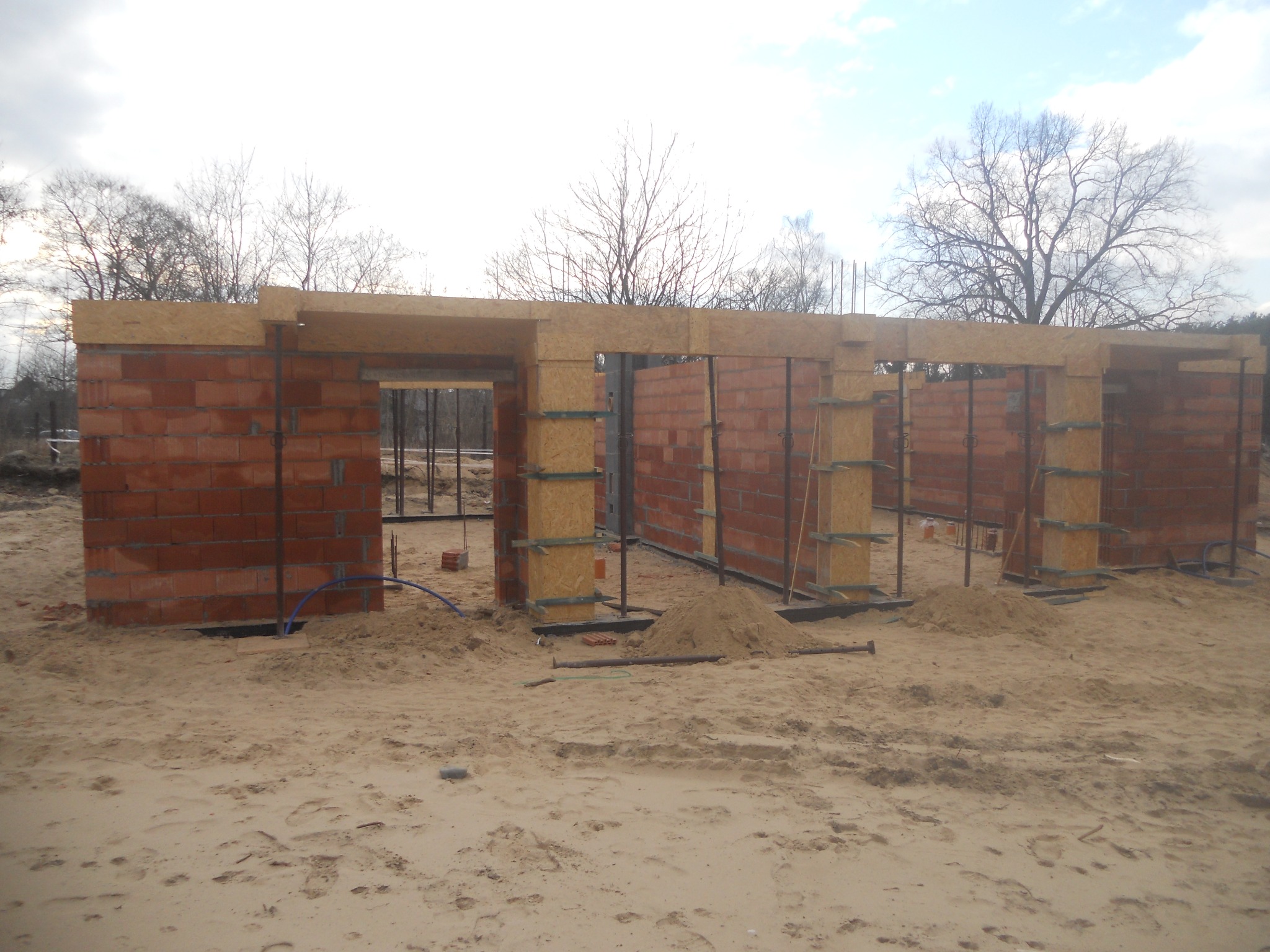 Wznoszenie murów z czerwonej cegły na fundamencie przyszłego domu, widoczne drewniane szalunki i metalowe pręty zbrojeniowe, piasek na placu budowy, drzewa w tle.