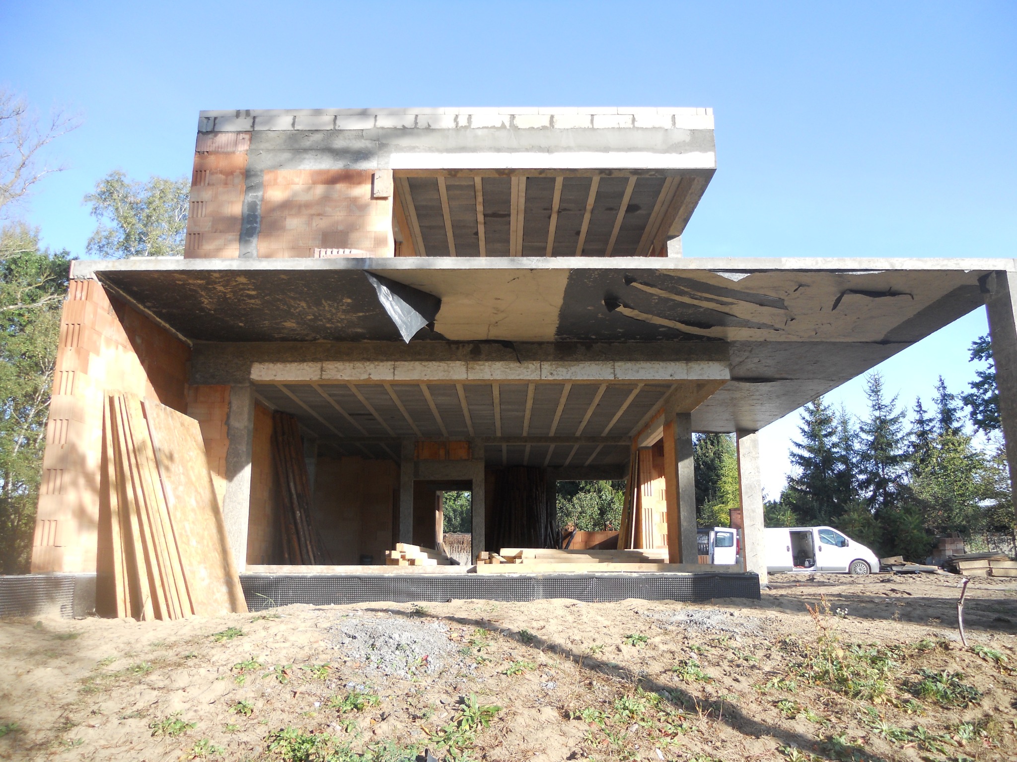 Surowy stan dwukondygnacyjnego domu murowanego z betonu i cegły, widoczne płyty betonowe stropu, niezabezpieczone folią, oraz biały samochód dostawczy w tle.