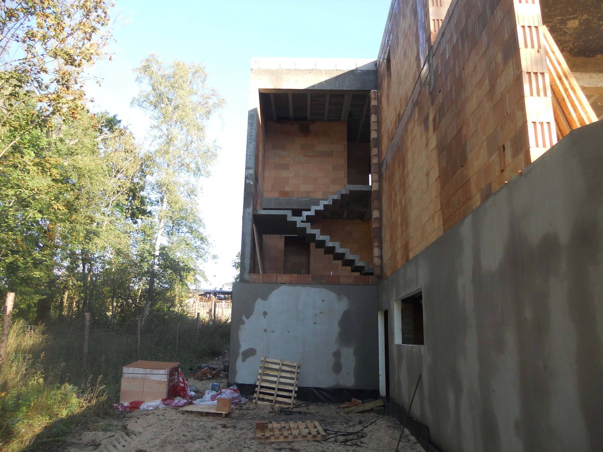 Surowy stan budynku w trakcie budowy z widocznymi schodami, ścianami z pustaków ceramicznych i otynkowaną ścianą zewnętrzną, palety leżą na ziemi, w tle drzewa.